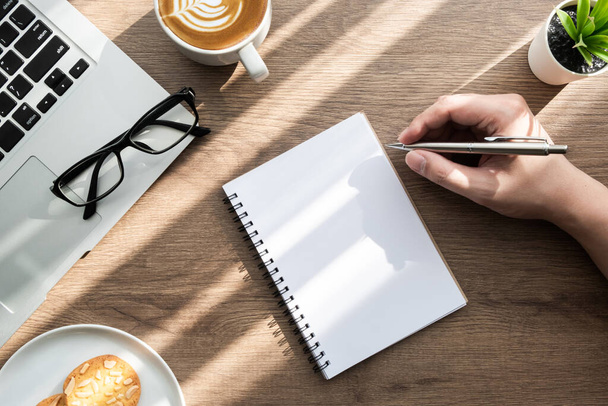 男は、クッキー、コーヒーや消耗品のカップと木製のオフィスの机のテーブルの上に空白のノートブックページに何かを書くつもりです。仕事のコンセプトでの朝の生活、トップビュー. - 写真・画像