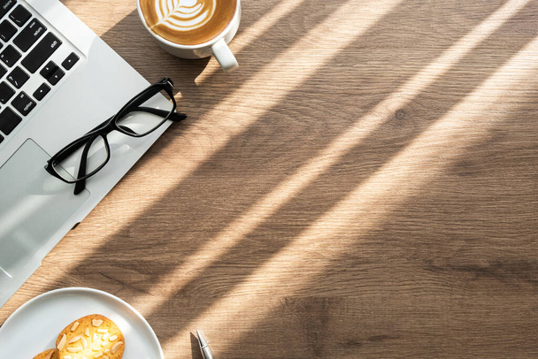 Draufsicht auf den Schreibtisch aus Holz mit einer Tasse Latte-Kaffee, Mandelkeksen und Bürobedarf mit Sonnenlicht. Das Arbeitsleben am Morgen. - Foto, Bild
