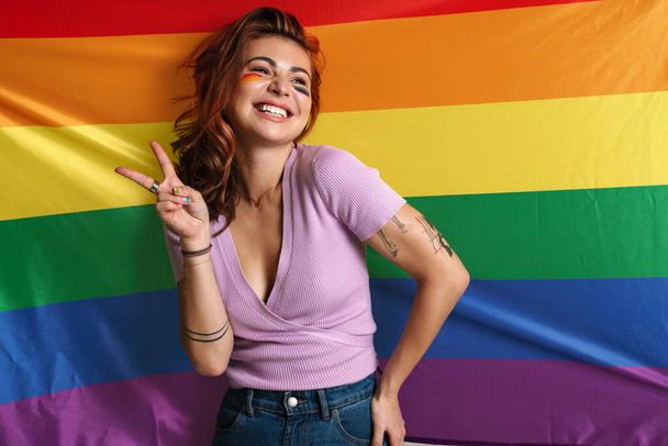 Cheerful young woman with makeup showing peace sign isolated over rainbow flag - Foto, imagen