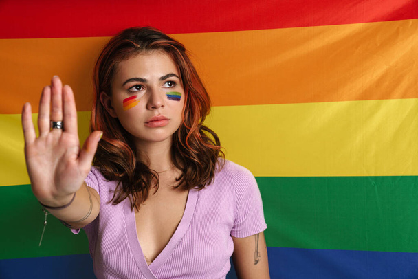 Displeased young woman with makeup showing stop gesture isolated over rainbow flag - Фото, изображение