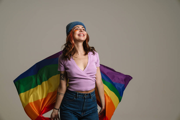 Happy beautiful woman smiling while posing with rainbow flag isolated over gray background - Foto, afbeelding