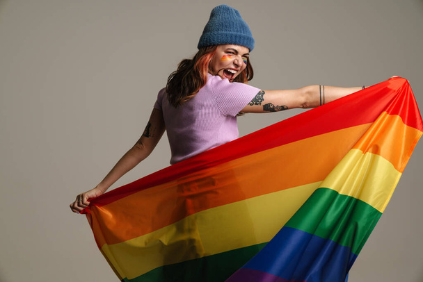 Cheerful beautiful woman laughing while posing with rainbow flag isolated over gray background - Foto, afbeelding