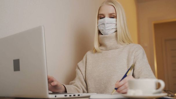 Jeune femme d'affaires en masque médical jetable tape sur le clavier de l'ordinateur portable et l'écriture. Coronavirus. - Photo, image