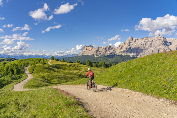 pretty active senior woman riding her electric mountain bike on the Pralongia Plateau in the Alta Badia Dolomites with awesome Sasso die Santa Cruce summit in Backg, South Tirol and Trentino, Italy - Photo, Image