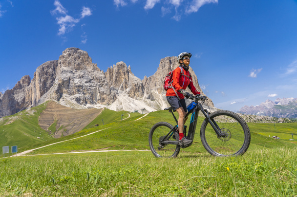 mulher sênior muito ativa montando sua bicicleta de montanha elétrica abaixo do famoso cume Sassolungo em Sella Dolomites de Selva Wolkenstein, Val Gardena, Tirol do Sul e Trentino, Itália - Foto, Imagem