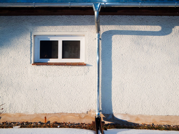 Detailansicht einer flachen Hausfassade mit Dachrinne, Fallrohr und kleines Fenster im seitlichen Abendlicht mit Schattenbild des Fallrohrs.  - Foto, immagini