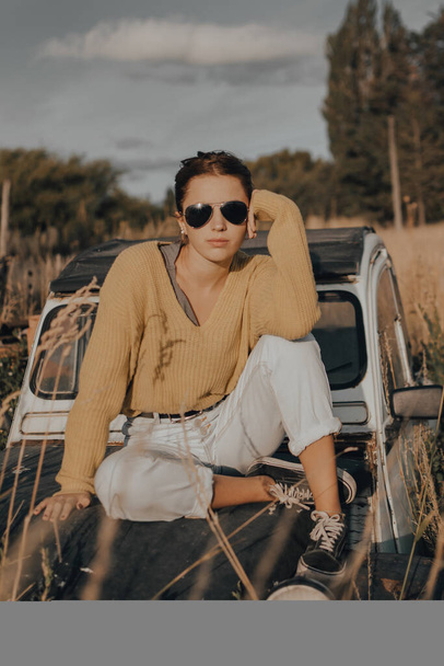 beautiful young girl looking at the camera sitting on an old car at sunset. - Photo, Image
