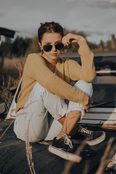 όμορφη νεαρή κοπέλα κοιτάζοντας την κάμερα κάθεται σε ένα παλιό αυτοκίνητο στο ηλιοβασίλεμα. - Φωτογραφία, εικόνα