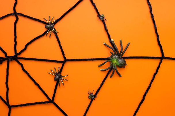 Many black toy spiders on artificial web on an orange background. Scary halloween flat lay composition on an orange background. Halloween decoration concept. - Фото, изображение
