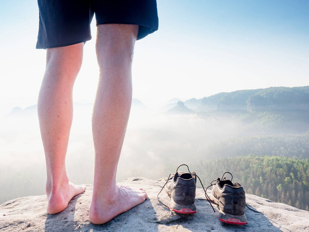Slim barefoot legs with hairy calves of runner stay at to removed sweaty running shoes on a rocky edge above a long deep vally in nature park. - Photo, Image