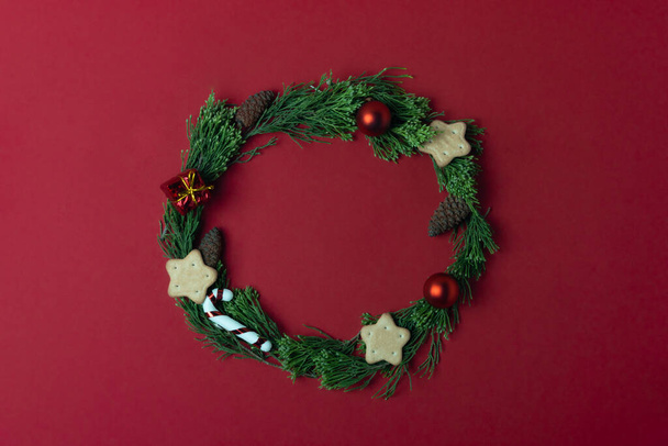 赤い背景にクリスマスのおもちゃやクッキーで飾られた針葉樹の枝の緑の花輪。クリスマスのコンセプト - 写真・画像