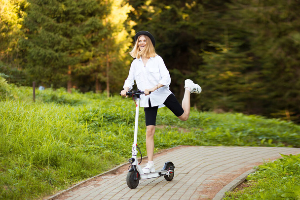 Chica hermosa alegre en sombrero negro y camisa blanca montando un scooter eléctrico en el parque de verano. Alquiler de vespa, libertad de movimiento - Foto, Imagen