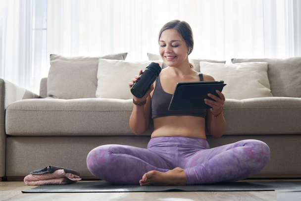 Beautiful Athletic Sporty Woman Sitting On Yoga Mat with tablet After Some Exercises and Drinks Protein Shake or water - Foto, imagen