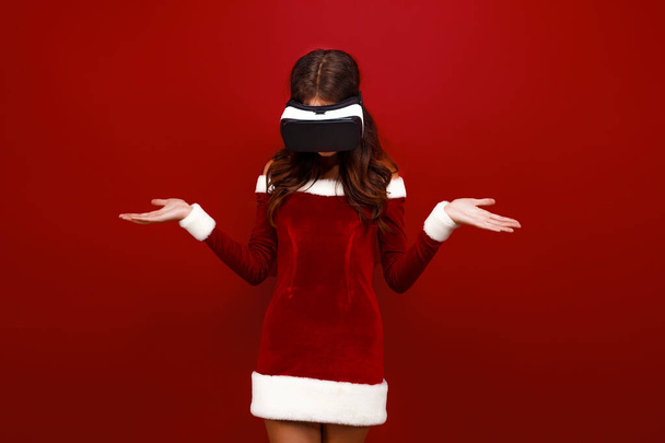 Erstaunte junge Weihnachtsmädchen im Weihnachtskleid suchen in Headset zeigt Hände zur Seite isoliert auf rotem Wandhintergrund.  - Foto, Bild