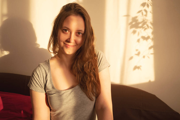 Young woman looking at camera in warm rays of sunset. Warm calm portrait of girl indoor. light and shadow on the face - Foto, imagen