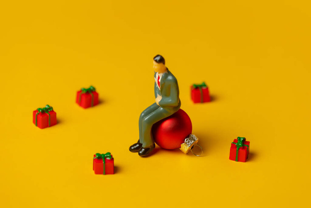 Miniature figure of man sits on Christmas bauble surrounded by gifts on yellow background - Photo, Image