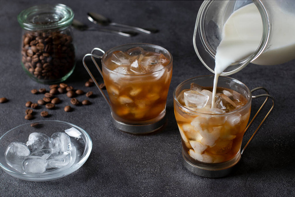 Zwei Tassen kalten Kaffees mit Eiswürfeln, in die aus einer Kanne Milch gegossen wird. Dunkler Hintergrund. Nahaufnahme. - Foto, Bild