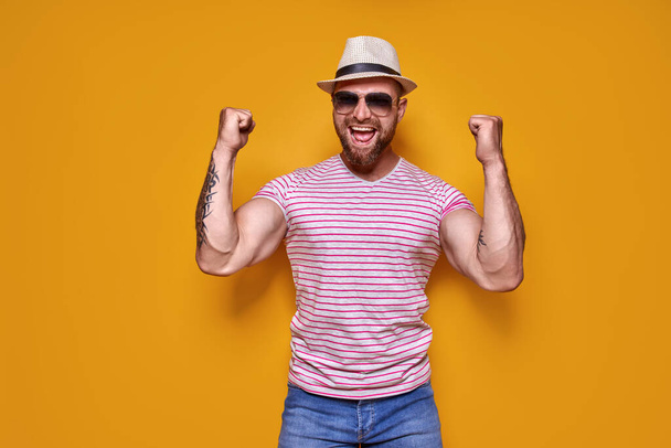 Image of excited bearded tattooed man in sunglasses and hat making winner gesture isolated yellow background. - Photo, image