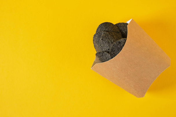 black potato chips in a package for french fries on a yellow background, copy space, eco friendly package - Fotoğraf, Görsel