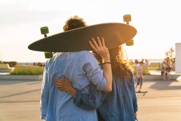 Romantic couple of skateboarders enjoy sunset embracing with longboard in hands in urban skate park - Photo, Image