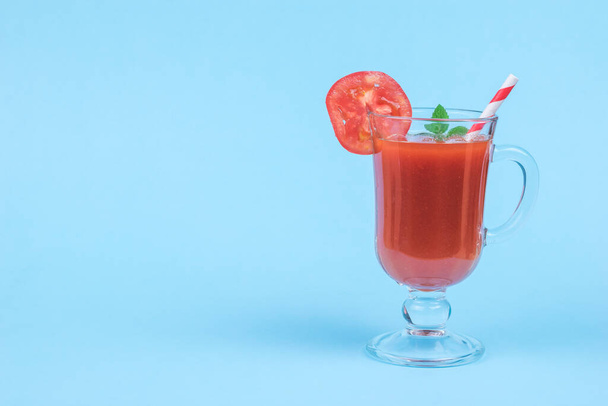 Glass glass with tomato juice, with a cocktail tube and mint leaves on a blue background. The concept of consumption of fresh vegetable juice. - Photo, Image