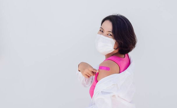 Έννοια εμβολιασμών. Εμβολιασμένη Ασιάτισσα γυναίκα με μάσκα προσώπου, ροζ αμάνικο δείχνοντας τον ροζ επίδεσμο στον ώμο της μετά τον εμβολιασμό, απομονωμένη σε λευκό χρώμα με αντιγραφικό χώρο. - Φωτογραφία, εικόνα