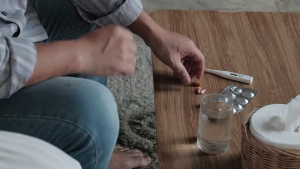 Asian man in a grey shirt and blue jeans sick leave because of illness takes temperature by a thermometer, takes medicine, and sleeps in blankets on sofa at home, tissues, water, and pills on table. - Imágenes, Vídeo
