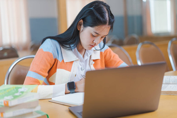Una studentessa universitaria asiatica stressante in uniforme studentesca che studia online, legge un libro, usa tablet o laptop digitali nella biblioteca universitaria nel tempo libero da sola. - Foto, immagini