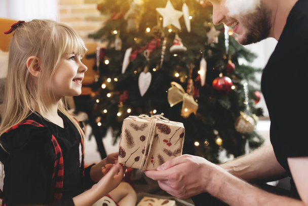 Kırmızı Noel Baba şapkalı genç baba süslü Noel ağacının yanındaki gülümseyen kızına Noel hediyesi veriyor. Kırmızı-siyah Noel kıyafeti giymiş bir kız. Gülümsüyorlar, yeni yıldan dolayı mutlular. - Fotoğraf, Görsel