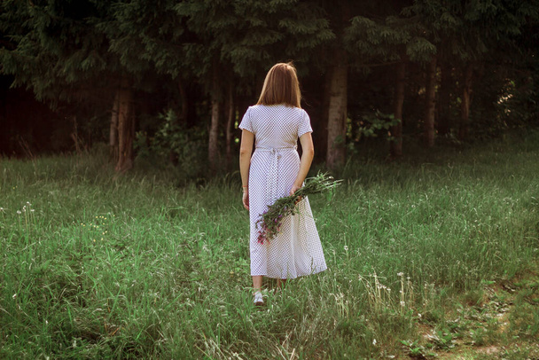A beautiful girl in a white dress stands with her back and holds a bouquet of wild flowers in her hands. Portrait of a slender girl with a bouquet of wild flowers in nature. Selective focus. - 写真・画像