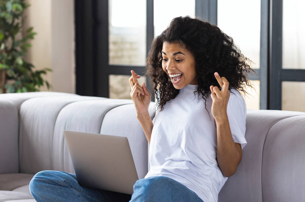 Nyertem a lottón. Izgatott fiatal afro-amerikai ujjakkal Keresztbe tett otthon ülve a kanapén nézi a laptop képernyőjén várja az eredményeket - Fotó, kép