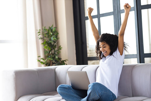 Excited young African American woman yelling yes, sitting at home on the couch looking at the laptop computer screen, celebrating victory, goal achievement raised arms up - Photo, image