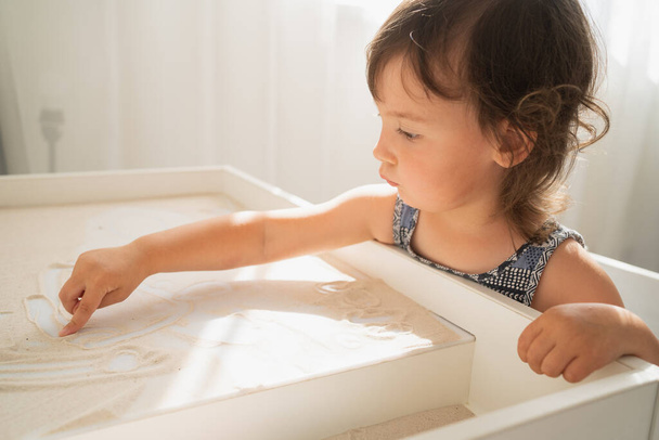 Mesa de dibujo de arena para niños. Una niña dibuja con el dedo sobre una mesa de arena clara. Desarrollo creativo en niños pequeños. - Foto, imagen