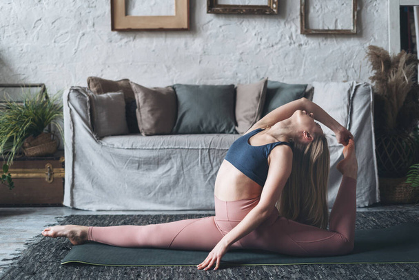 Jonge sportieve vrouw die thuis yoga beoefent, fitte vrouw zittend in One Legged King Pigeon poseren in de woonkamer op de vloer in de buurt van een grijze bank, het dragen van sport beha en roze leggins. Actieve levensstijl - Foto, afbeelding
