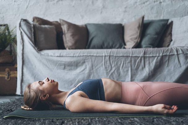 Giovane donna rilassata in abbigliamento attivo sdraiata in posa Dead Body durante la pratica dello yoga a casa, femminile facendo Savasana o posa cadavere in soggiorno moderno con divano. Concetto di consapevolezza e riduzione dello stress - Foto, immagini