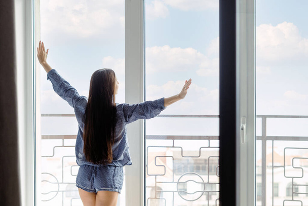 Młoda kobieta ubrana w piżamę z pięknymi długimi włosami rozciągającymi ramiona lub ćwicząca gimnastykę po przebudzeniu i wyglądająca przez okno panoramiczne w domu, ciesząca się niesamowitym widokiem na miasto rano w domu - Zdjęcie, obraz