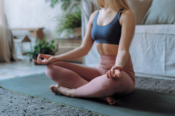 Обрезанный снимок активной женщины в спортивной одежде, сидящей в позе медитации лотоса и держащейся за руки жестом мудры во время занятий йогой и медитацией в гостиной дома, избирательный фокус - Фото, изображение