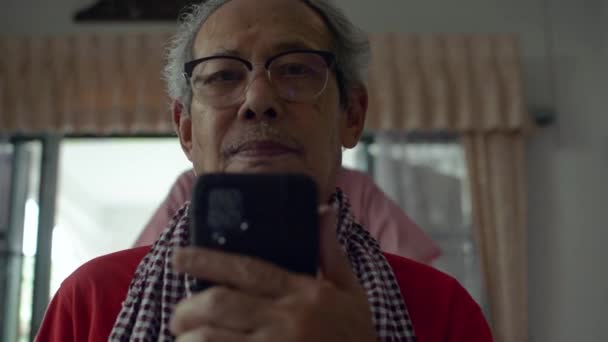 Ázsiai végzős ősz haj nagyapa néz TV-t mobil okostelefon a nappaliban, míg az imádnivaló unokája megközelíteni, és megölelni hátulról boldogan. Kötődési kapcsolat két generációs családban. - Felvétel, videó
