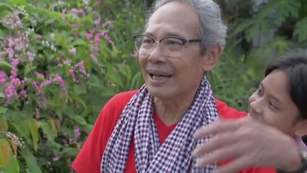 Cheerful Asian teen granddaughter approach and hug her healthy senior gray hair grandfather in eyeglasses who caring the flowers in the backyard garden. Bonding in two generation of a family. - Materiaali, video