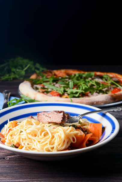 Closeup of rustic spaghetti with beefsteak, vegetables, garlic and fresh rosemary. Italian food. Italian cuisine. Concept for a tasty and hearty dish. Over dark wooden background with pizza. - Foto, Imagem