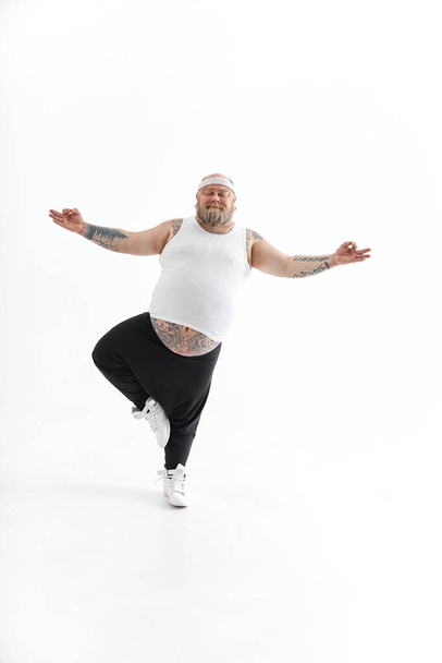 Щасливий товстий чоловік з великим животом і татуюваннями в спортивному одязі позує на білому тлі
 - Фото, зображення