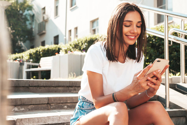 Portrait of young beautiful smiling hipster woman in trendy summer jeans shorts. Sexy carefree model posing in the street. Sitting at the stairs.Looking at smartphone screen, using phone apps - Photo, image