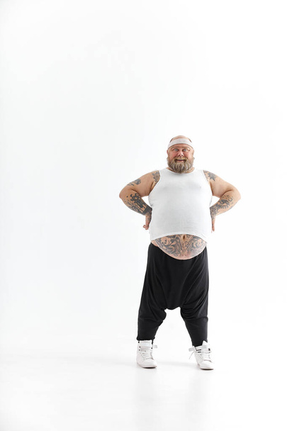 Hombre gordo feliz con gran vientre y tatuajes en ropa deportiva posando sobre fondo blanco - Foto, Imagen