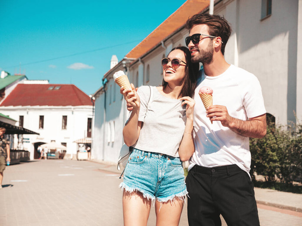 Smiling beautiful woman and her handsome boyfriend. Woman in casual summer clothes. Happy cheerful family. Couple posing on the street background in sunglasses. Eating tasty ice cream in waffles cone - Фото, изображение