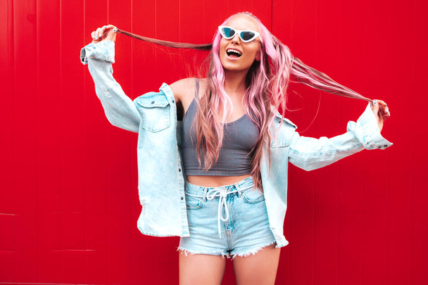 Young beautiful smiling hipster woman in trendy summer jeans shorts with pink hairstyle. Sexy carefree model posing in the street near red wall. Positive model outdoors in sunglasses - Foto, Imagen