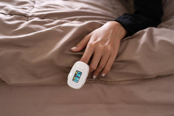 Hand mit Pulsoximeter oder Blutsauerstoffmessgerät auf dem Bett zu Hause isoliert. Diagnose Selbstkontrolle. Medizinische und medizinische Ausrüstung.Coronavirus covid-19 Ausbruch - Foto, Bild