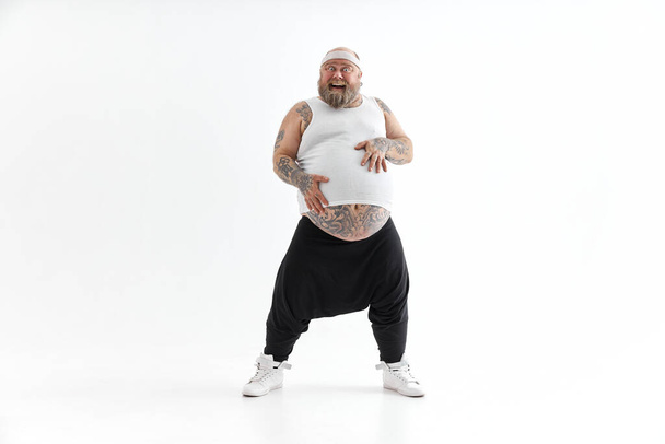 Χαρούμενος χοντρός με μεγάλη κοιλιά και τατουάζ στα αθλητικά φοράει το στομάχι του. - Φωτογραφία, εικόνα