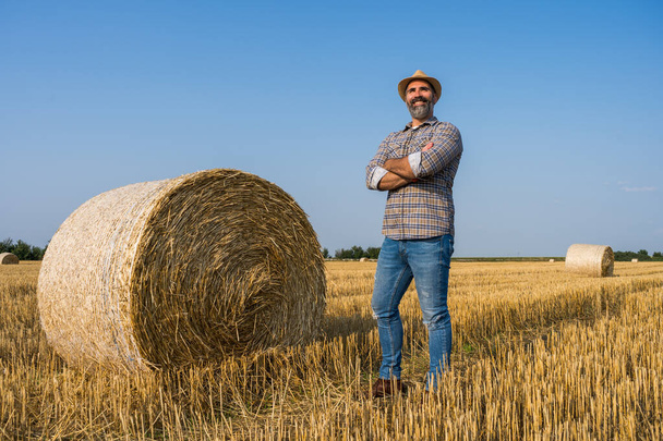 Χαρούμενος αγρότης στέκεται δίπλα σε δεμάτια άχυρου. Είναι ικανοποιημένος λόγω της επιτυχημένης συγκομιδής. - Φωτογραφία, εικόνα
