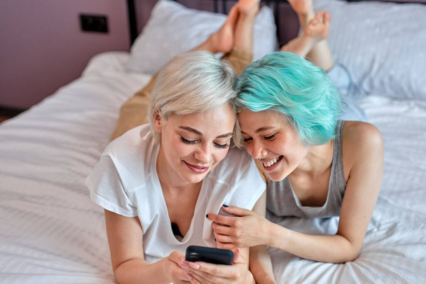 Positive lesbian couple having sun on bed using smartphone, watching video - Фото, изображение