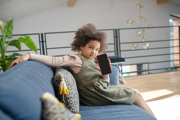Little girl showing smartphone screen sitting on sofa - Foto, Imagen
